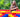 A young girl smiles while standing beside a colorful structure made of large building blocks in a vibrant, outdoor setting.