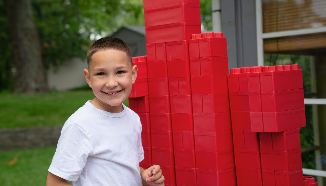 A smiling kid happily playing with red jumbo blocks from Biggo Blocks outdoors.