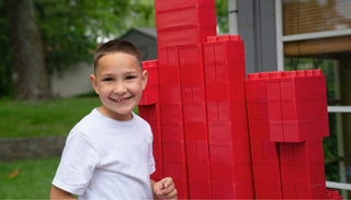 A smiling kid happily playing with red jumbo blocks from Biggo Blocks outdoors.