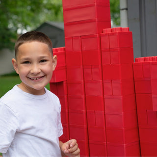 A smiling kid happily playing with red jumbo blocks from Biggo Blocks outdoors.