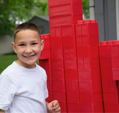 A smiling kid happily playing with red jumbo blocks from Biggo Blocks outdoors.