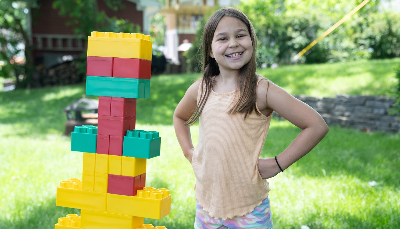 A young girl proudly stands next to a colorful structure built with Biggo Blocks in a sunny backyard.