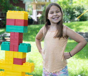 A young girl proudly stands next to a colorful structure built with Biggo Blocks in a sunny backyard.