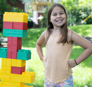 A young girl proudly stands next to a colorful structure built with Biggo Blocks in a sunny backyard.