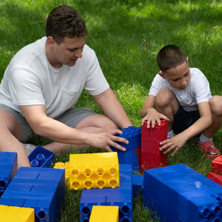 Father and son playing together on the grass with colorful jumbo building blocks, enjoying outdoor bonding time on a sunny day.