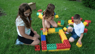 Three children building with colorful jumbo building blocks on green grass. 