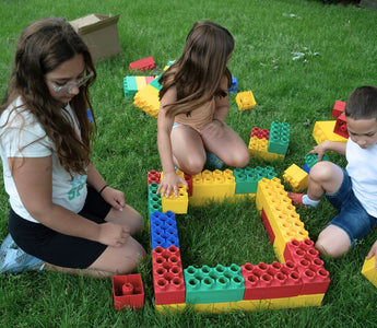 Three children building with colorful jumbo building blocks on green grass. 
