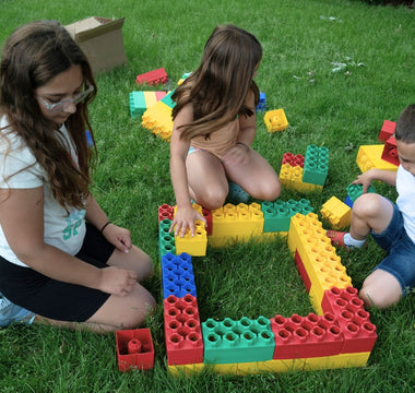 Three children building with colorful jumbo building blocks on green grass. 