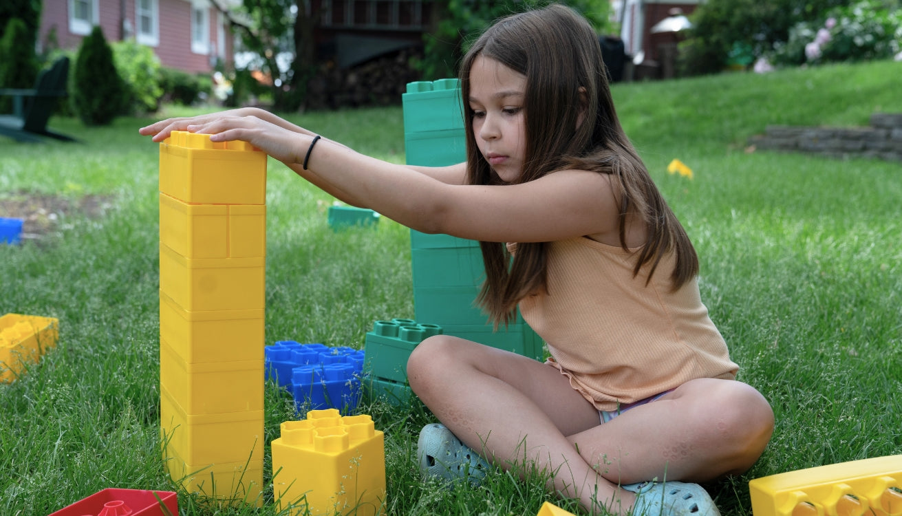 A girl building with colorful blocks on the grass, focused on stacking a yellow block. 