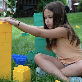 A girl building with colorful blocks on the grass, focused on stacking a yellow block. 