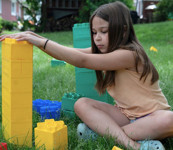A girl building with colorful blocks on the grass, focused on stacking a yellow block. 