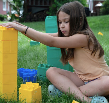 A girl building with colorful blocks on the grass, focused on stacking a yellow block. 