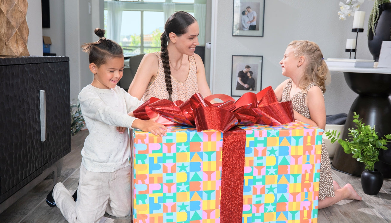 Two children and a woman excitedly unwrapping a big birthday gift with colorful wrapping and bows. 