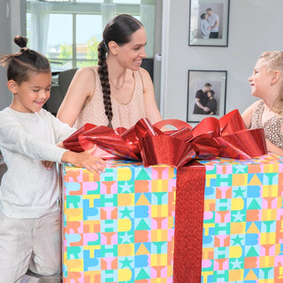 Two children and a woman excitedly unwrapping a big birthday gift with colorful wrapping and bows. 