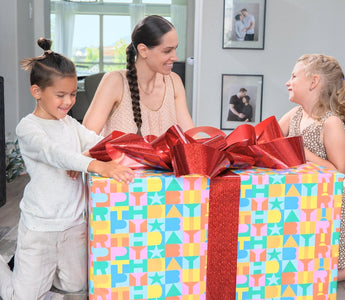Two children and a woman excitedly unwrapping a big birthday gift with colorful wrapping and bows. 