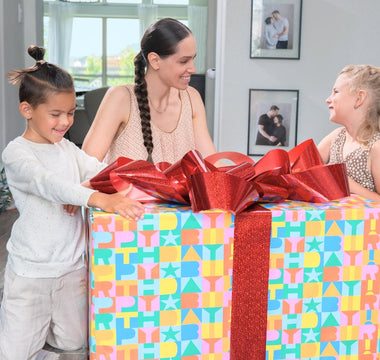 Two children and a woman excitedly unwrapping a big birthday gift with colorful wrapping and bows. 