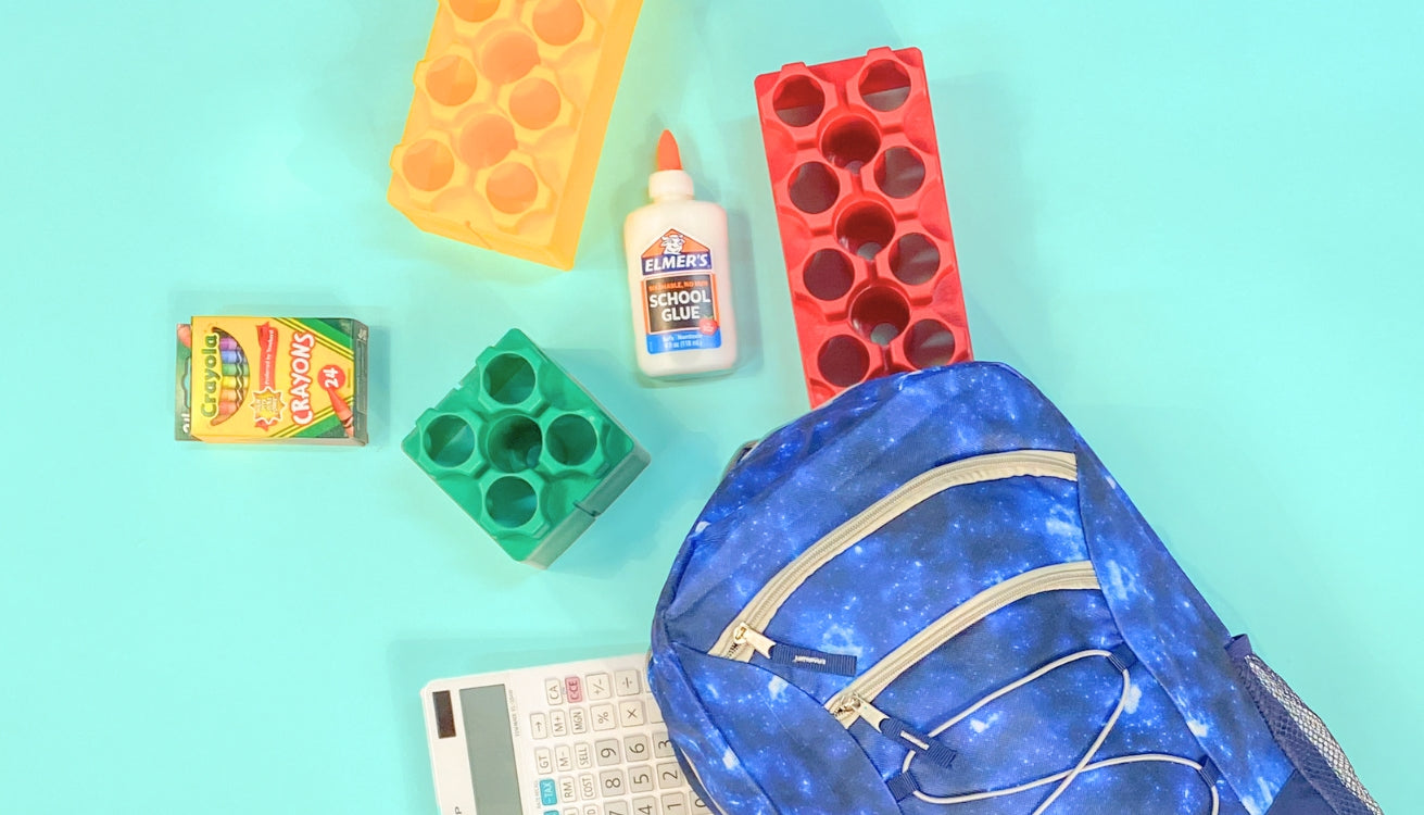 A back-to-school setup featuring a blue galaxy-themed backpack, colorful Biggo Blocks, a box of Crayola crayons, a bottle of Elmer's school glue, and a calculator, all laid out on a bright turquoise background.