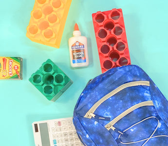 A back-to-school setup featuring a blue galaxy-themed backpack, colorful Biggo Blocks, a box of Crayola crayons, a bottle of Elmer's school glue, and a calculator, all laid out on a bright turquoise background.