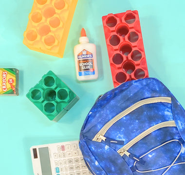 A back-to-school setup featuring a blue galaxy-themed backpack, colorful Biggo Blocks, a box of Crayola crayons, a bottle of Elmer's school glue, and a calculator, all laid out on a bright turquoise background.