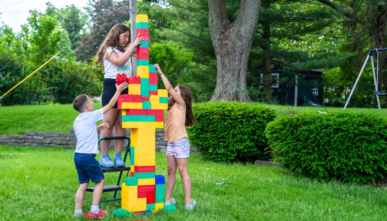 Three children working together to build a tall tower using colorful extra-large building blocks in a backyard setting.