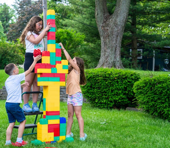 Three children working together to build a tall tower using colorful extra-large building blocks in a backyard setting.