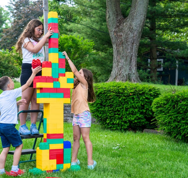 Three children working together to build a tall tower using colorful extra-large building blocks in a backyard setting.