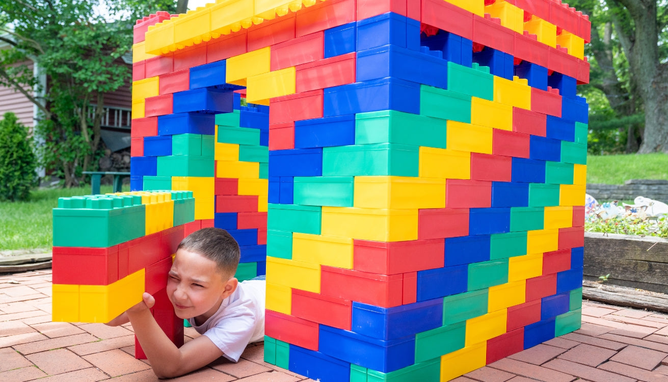 A young boy peeks out from behind a colorful building made of Biggo Blocks, enjoying a playful moment surrounded by the extra-large building blocks.