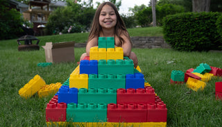 Smiling young girl building a colorful pyramid with oversized Biggo Blocks on a lush green lawn, demonstrating the joy and creativity inspired by sensory play outdoors.
