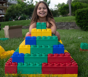 Smiling young girl building a colorful pyramid with oversized Biggo Blocks on a lush green lawn, demonstrating the joy and creativity inspired by sensory play outdoors.