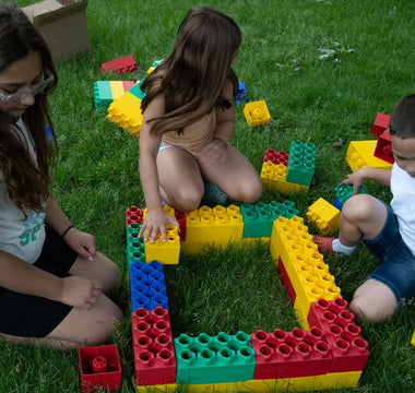 Three children sitting on grass, collaboratively arranging jumbo red, yellow, green, and blue blocks into a large square frame.