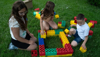 Three children sitting on grass, collaboratively arranging jumbo red, yellow, green, and blue blocks into a large square frame.