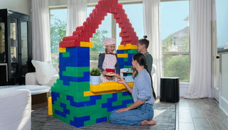 A mother and her two children engage in imaginative play at home, using large Biggo Blocks to construct a colorful playhouse or shop. One child, dressed as a chef, pretends to serve food through a window, creating a playful and creative moment.