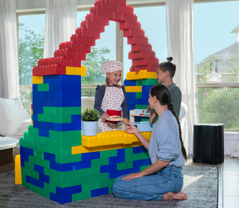 A mother and her two children engage in imaginative play at home, using large Biggo Blocks to construct a colorful playhouse or shop. One child, dressed as a chef, pretends to serve food through a window, creating a playful and creative moment.