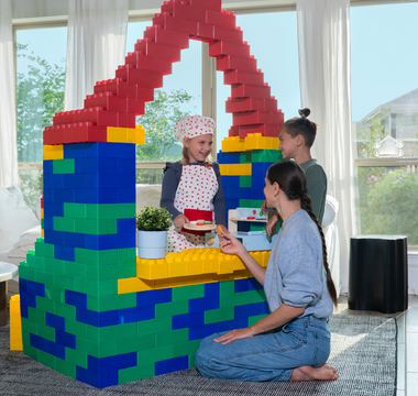 A mother and her two children engage in imaginative play at home, using large Biggo Blocks to construct a colorful playhouse or shop. One child, dressed as a chef, pretends to serve food through a window, creating a playful and creative moment.