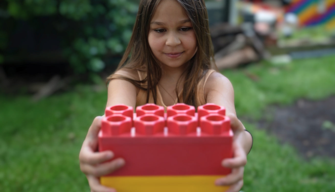 A young girl holding a large red and yellow building block with a focused expression, enjoying creative play in a backyard setting.