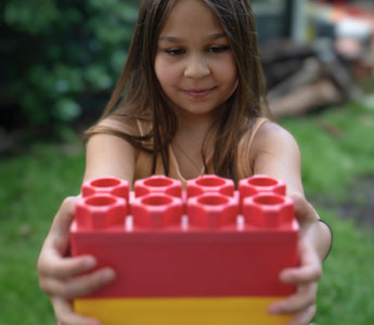 A young girl holding a large red and yellow building block with a focused expression, enjoying creative play in a backyard setting.