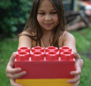 A young girl holding a large red and yellow building block with a focused expression, enjoying creative play in a backyard setting.