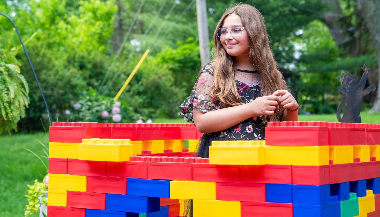 A young girl smiles while standing beside a colorful structure made of large building blocks in a vibrant, outdoor setting.