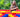 A young girl smiles while standing beside a colorful structure made of large building blocks in a vibrant, outdoor setting.