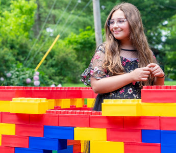 A young girl smiles while standing beside a colorful structure made of large building blocks in a vibrant, outdoor setting.