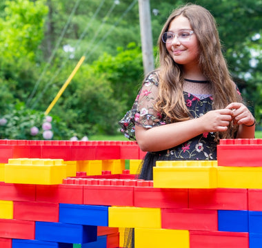 A young girl smiles while standing beside a colorful structure made of large building blocks in a vibrant, outdoor setting.