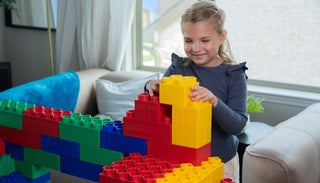 A young girl happily builds a colorful structure with large red, green, blue, and yellow building blocks in a cozy living room setting.