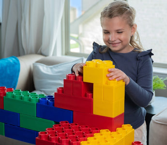 A young girl happily builds a colorful structure with large red, green, blue, and yellow building blocks in a cozy living room setting.