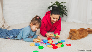 A mother and her young daughter playing with colorful building blocks on the carpet, while a ginger cat watches nearby.