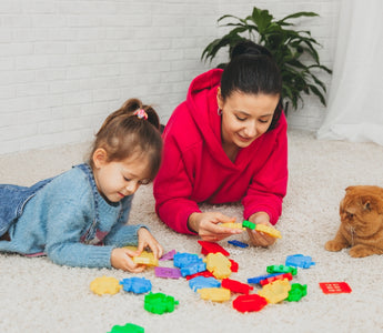 A mother and her young daughter playing with colorful building blocks on the carpet, while a ginger cat watches nearby.