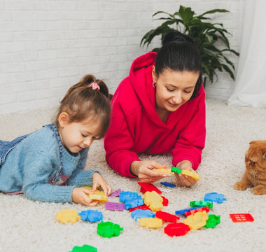 A mother and her young daughter playing with colorful building blocks on the carpet, while a ginger cat watches nearby.