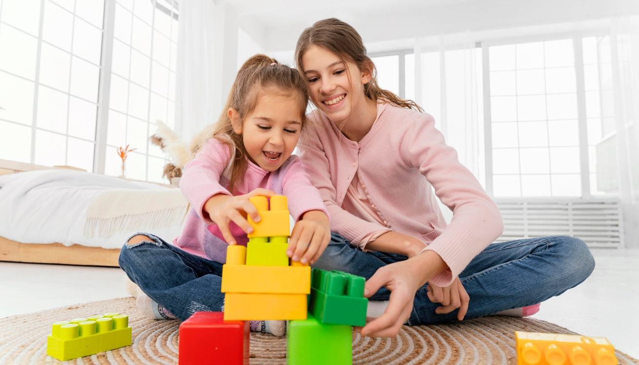 Two young girls playing together with colorful Biggo Blocks, building a tower on the floor in a bright, sunlit room.