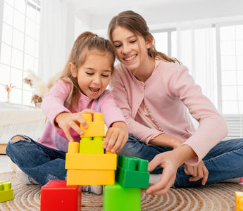 Two young girls playing together with colorful Biggo Blocks, building a tower on the floor in a bright, sunlit room.