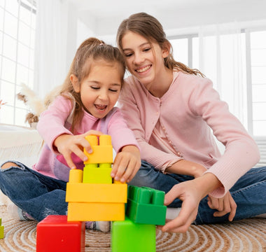 Two young girls playing together with colorful Biggo Blocks, building a tower on the floor in a bright, sunlit room.