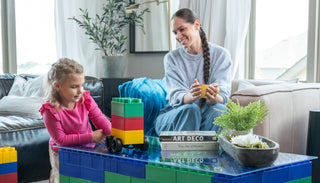 A young girl playing with Biggo Blocks, assembling a vehicle while her mother watches and smiles from the couch, showcasing a creative and playful moment with extra large building blocks.